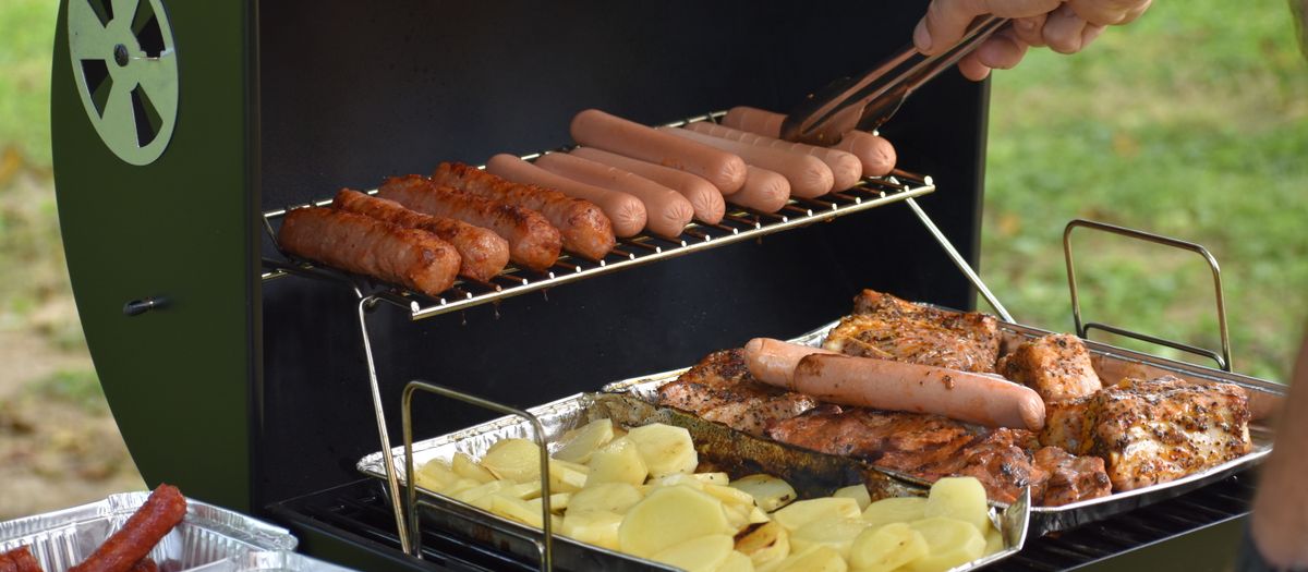 Outdoor barbecue with hotdogs, sausages, potatoes, and chicken grilling on a sunny day.