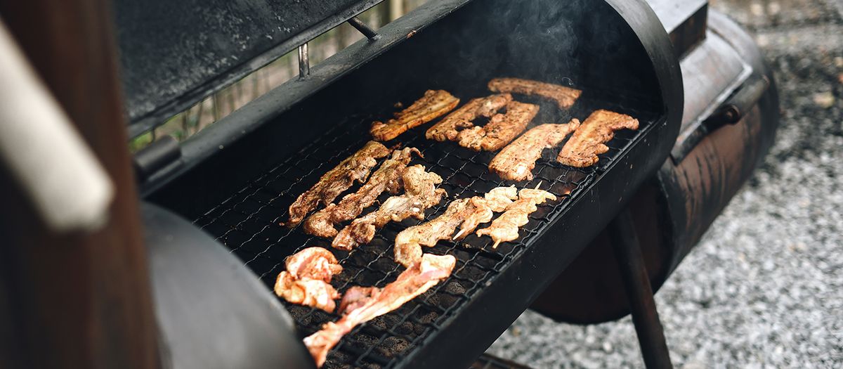 Close-up of a barbecue grill outdoors with various meat strips cooking, exuding rich smoke.